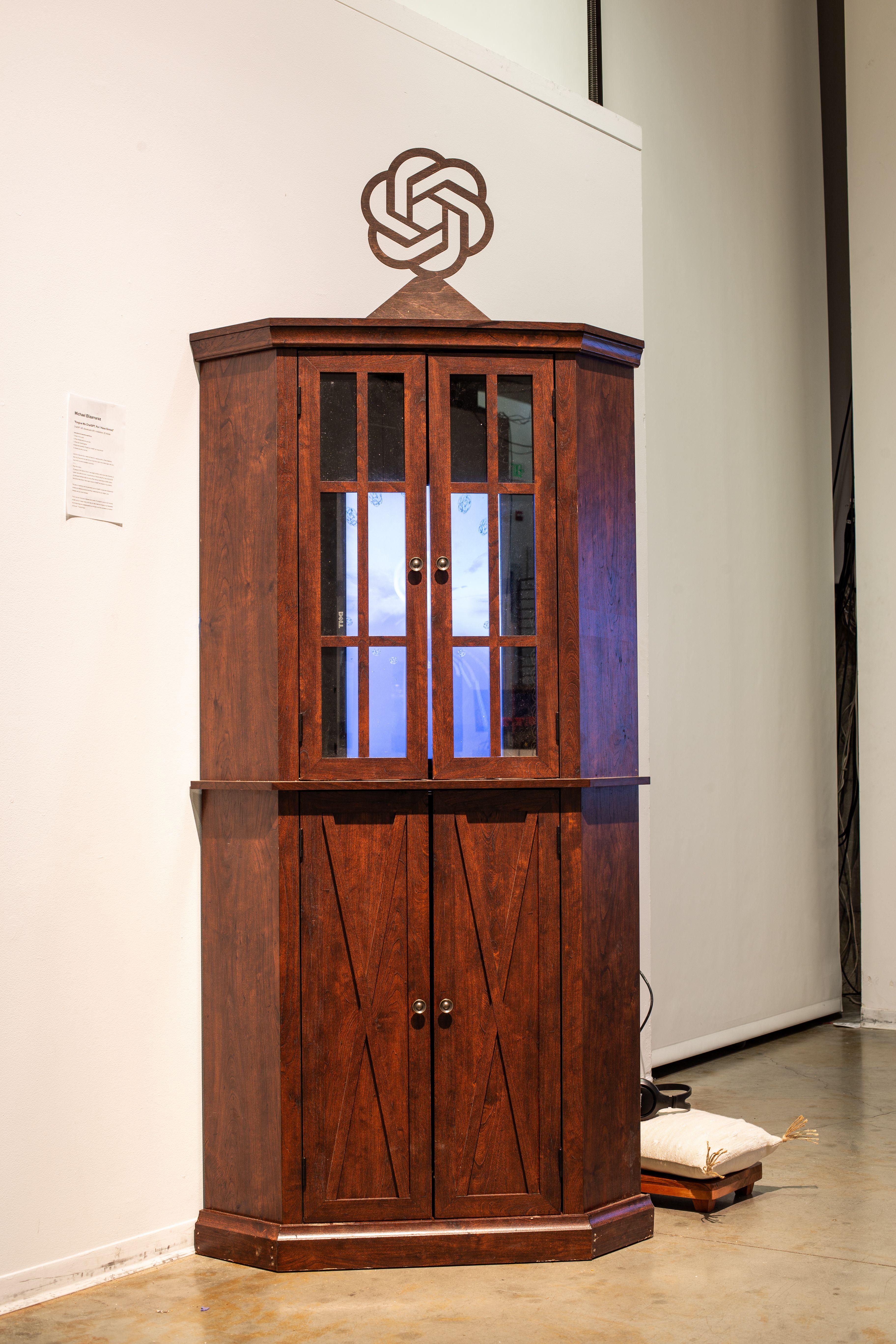 A large wooden cabinet with windowed doors, within which a vertically oriented television play a video of swirling, organic forms. On top of the cabinet is a wooden cutout of the ChatGPT logo, a geometric figure that resembles a hexagon, but has rounded a