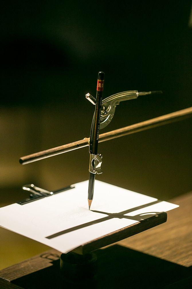 This is a close up of a pencil and white paper pad. The pencil is held by a metal tool that is connected to a long, thin wooden rod. There seems to be nothing drawn on the paper.