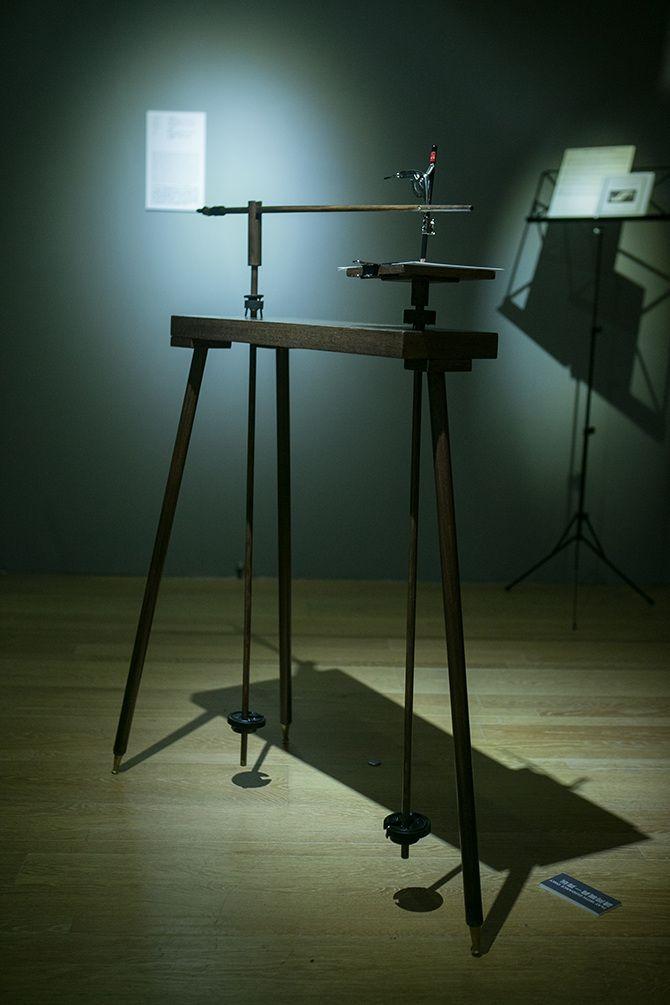 This is a close up picture of a device with three thin black legs and two long stick-like items that look like ski poles not long enough to touch the ground attached to it.
