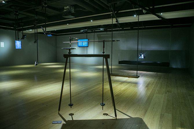 This is an angled photo of a device with three thin black legs (one on one side, two on the other) and two long stick-like items that look like ski poles not long enough to touch the ground attached to it. In the background, there is a swing, and behind t