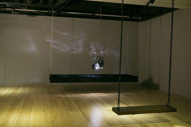 This is an angled photo of a stage light that is relatively close to the ground pointing down. The stage light has a rectangular box of black material suspended in the air around it. There are some abstract light reflections on the wall. In the right half