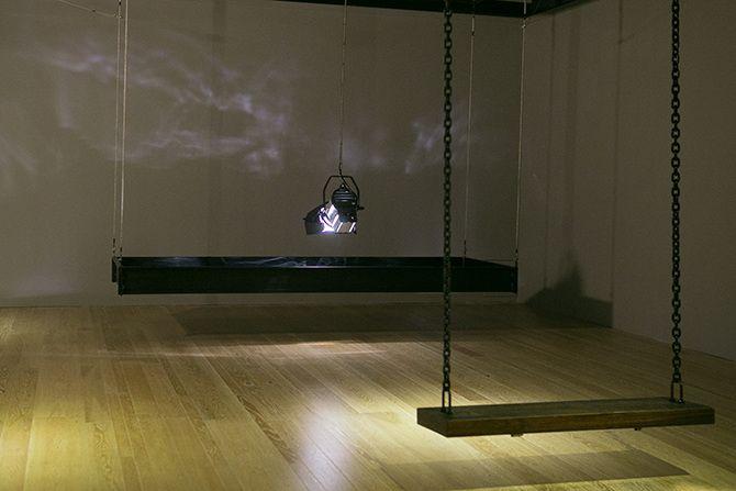 This is an angled photo of a stage light that is relatively close to the ground pointing down. The stage light has a rectangular box of black material suspended in the air around it. There are some abstract light reflections on the wall.
