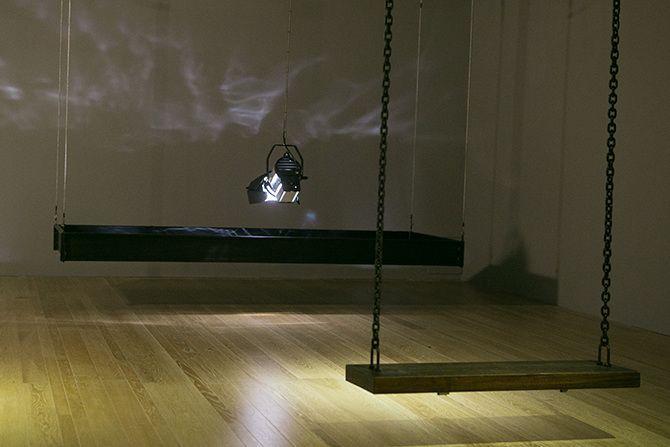 This is an angled photo of a stage light that is relatively close to the ground pointing down. The stage light has a rectangular box of black material suspended in the air around it. There are some abstract light reflections on the wall. In the right half