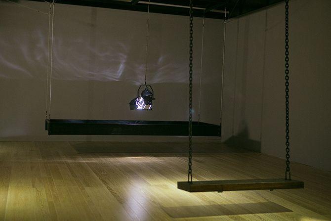 This is an angled photo of a stage light that is relatively close to the ground pointing down. The stage light has a rectangular box of black material suspended in the air around it. There are some abstract light reflections on the wall. In the right half