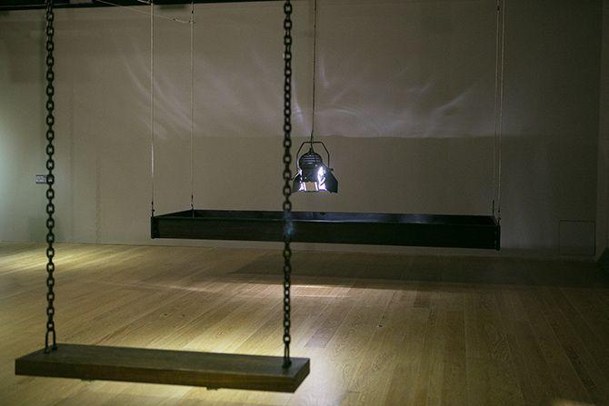 This is an angled photo of a stage light that is relatively close to the ground pointing down. The stage light has a rectangular box of black material suspended in the air around it. There are some abstract light reflections on the wall. In the left half 