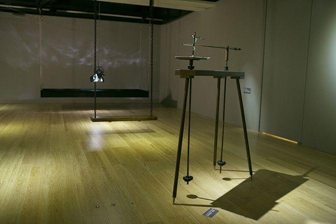 This is an angled photo of a device with three thin black legs and two long stick-like items that look like ski poles not long enough to touch the ground attached to it. In the background, there is a swing, and behind that, there is a stage light facing d