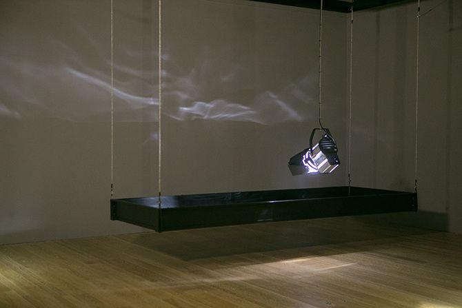 This is a photo of a stage light that is relatively close to the ground pointing down. The stage light has a rectangular box of black material suspended in the air around it. There are some abstract light reflections on the wall.