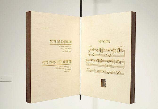 There is a plain beige wooden block shaped in the form of an open book suspended in air. The wood has brown inscriptions. The left block has the inscriptions "NOTE DE L'AUTER" underlined with text too small to read under it, then "NOTE FROM THE AUTHOR" un