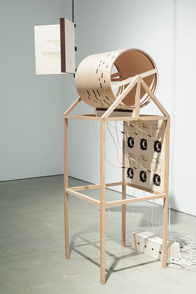 This is a diagonal view of the light beige machine and book suspended in the air. The machine is supported by thin wooden beams and has a cylindrical figure and six speakers, all with lines coming out of them. The text in the book is too small and faint t