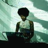 A dark-skinned woman stands behind a laptop in a dimly lit room, performing live coding. She has a short afro and is dressed in a sleeveless white top. Her face and body are illuminated by projected code, with lines of text and symbols wrapping around her