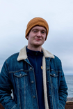 A blonde-haired Latvian-American man wearing a orange beanie against an ocean backdrop. His hands are in the pockets of his jacket.