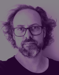 Close-up of the face of a white man with curly hair and a short salt and pepper beard. He is wearing glasses and a black t-shirt. The image is tinted purple.