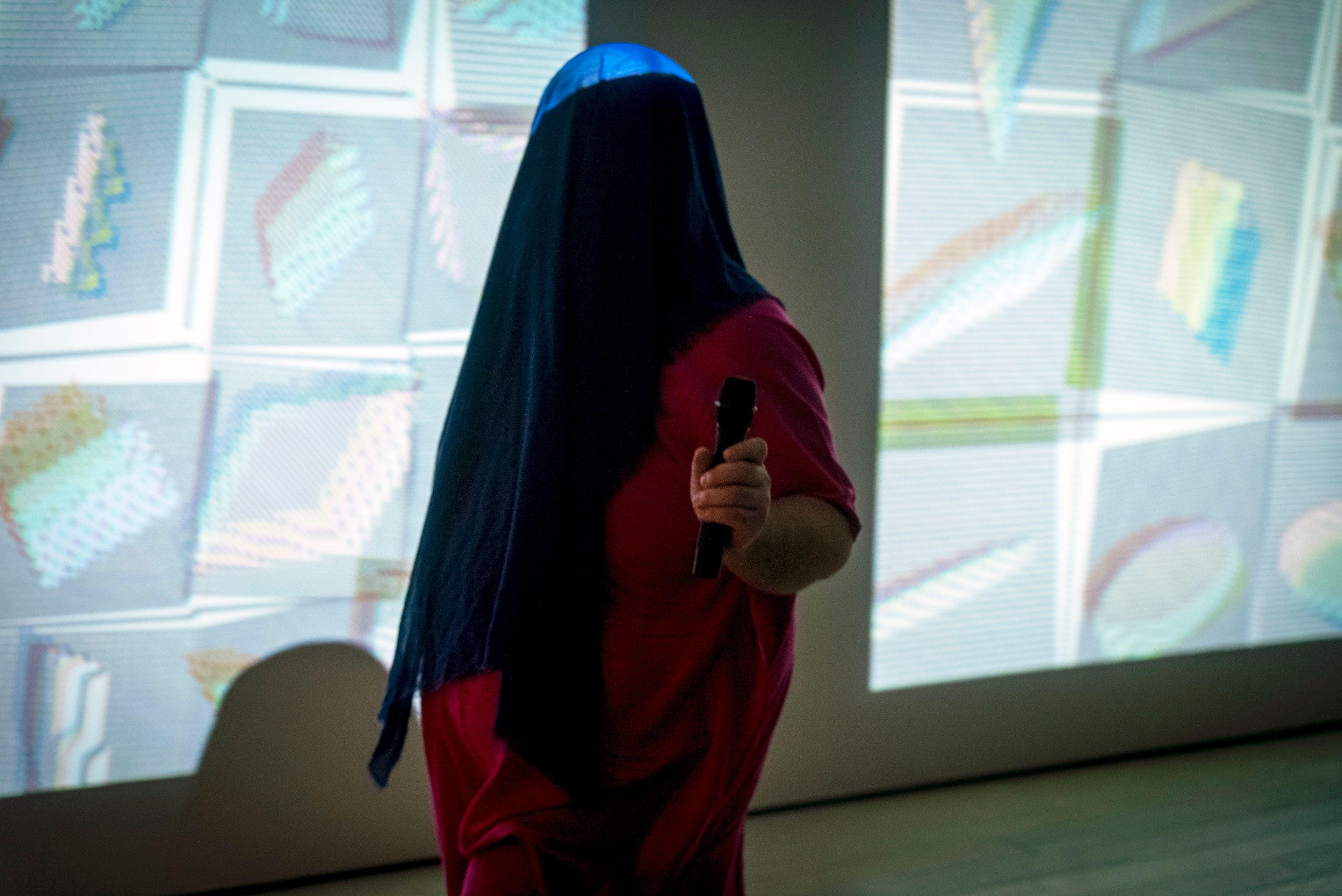 edgar in pink dress and blue head covering holds a mic and walks in front of projected graphic solids