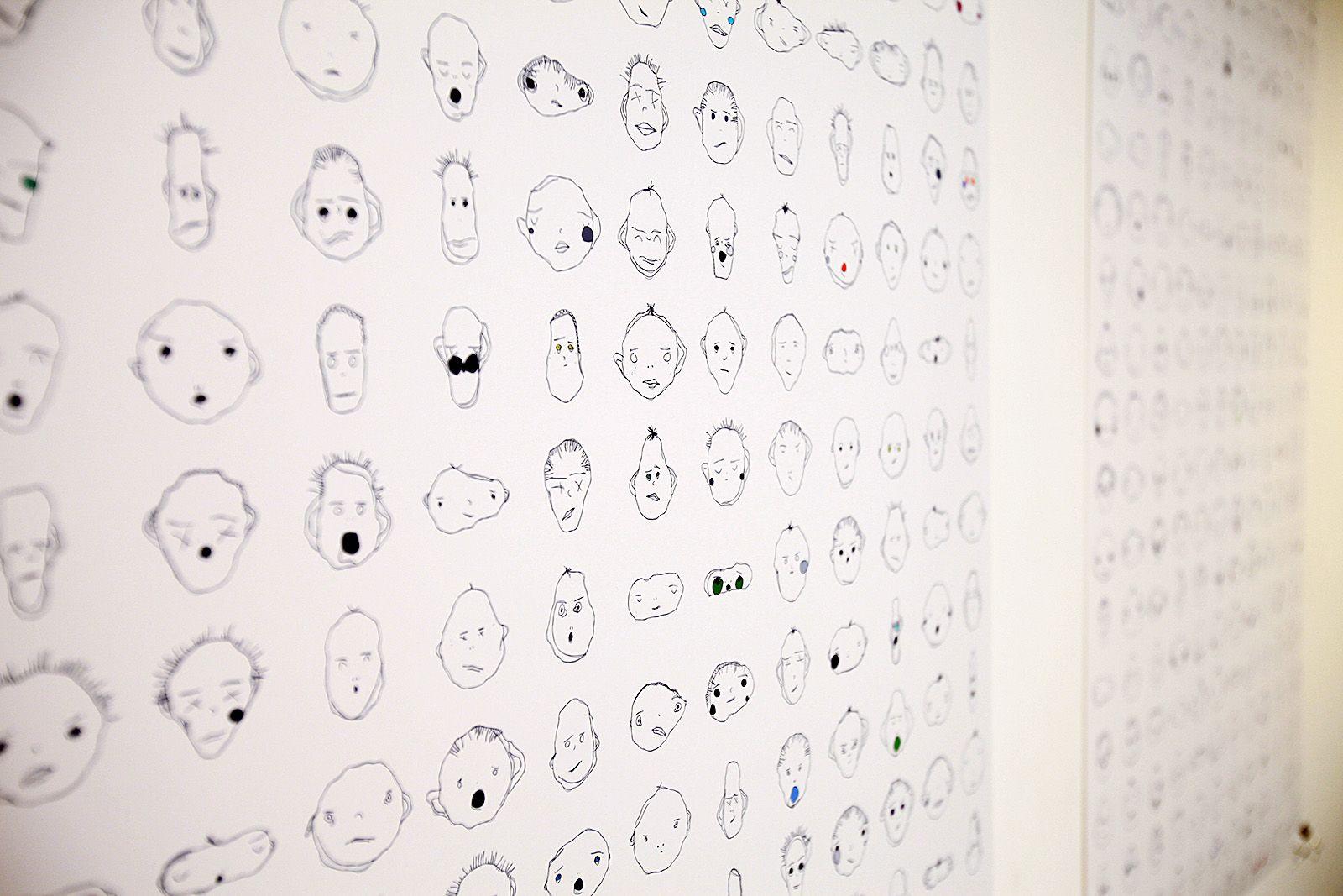 This is a diagonal close up view of two prints of various equally spaced apart weird faces. All of them have two eyes, two eyebrows, and a mouth. Some have hair, a nose, ears, or blush. All have unique expressions and are line drawn in black. Almost none 