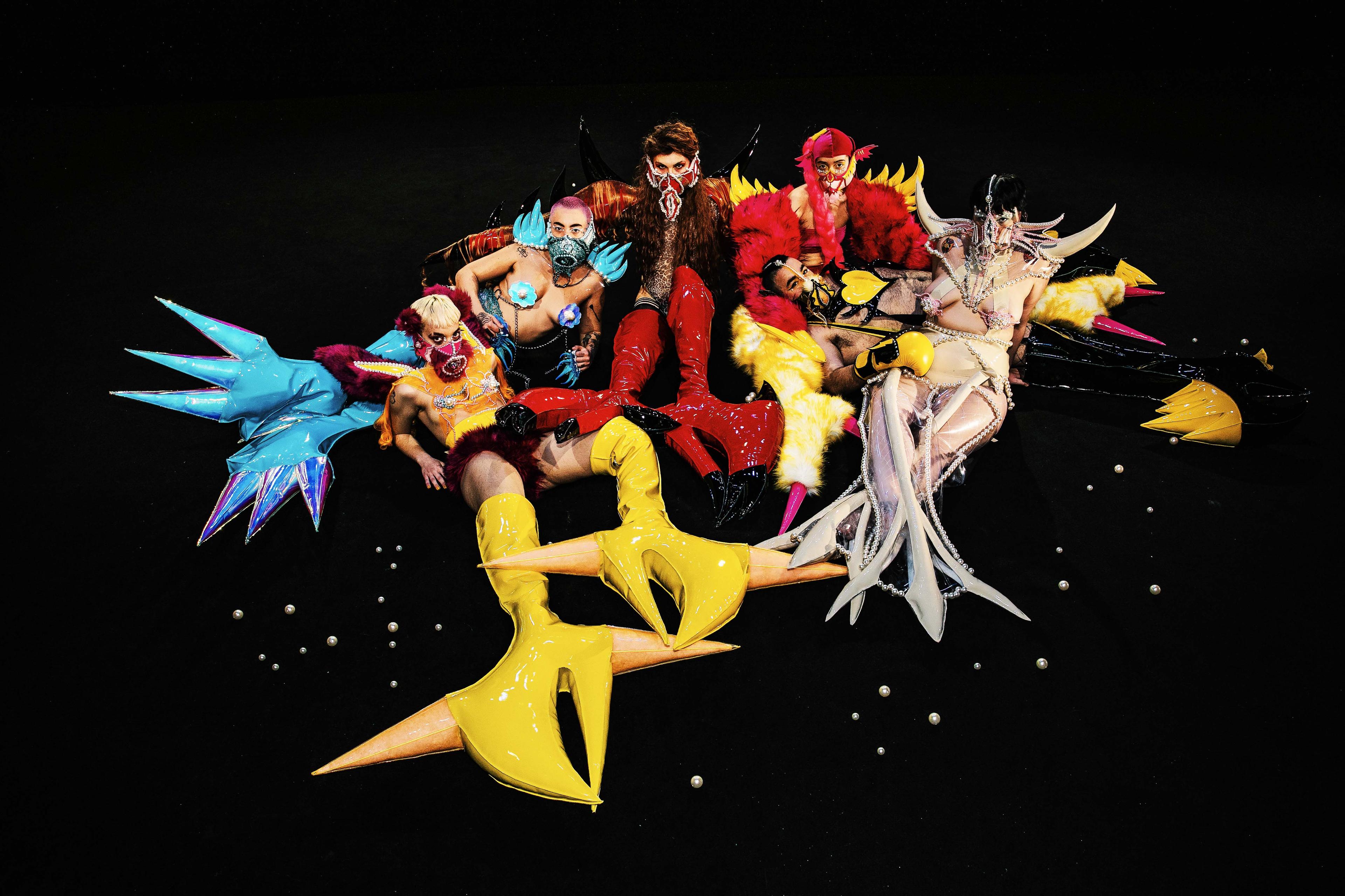 Six human-like creatures who look like superheroes are lying close to each other on a black background. Each creature is wearing a vibrant coloured vinyl bodysuit with knee-high vinyl boots with large platforms which elongate the limbs, fur details, shiny