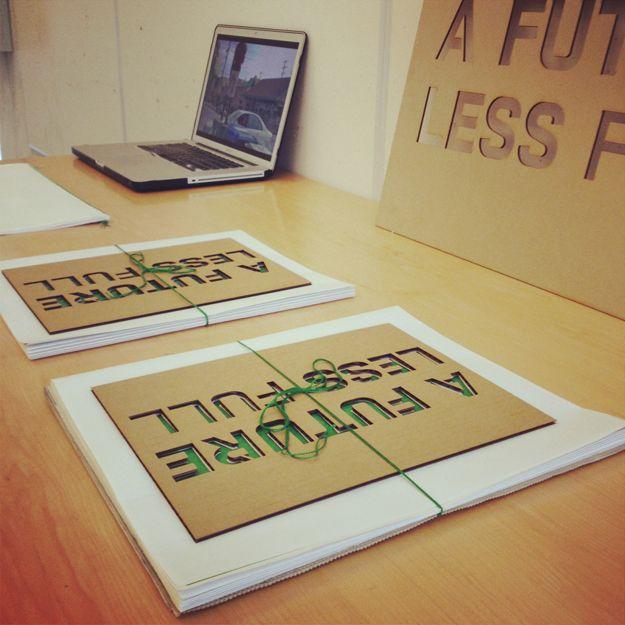 There are two stacks of paper on a desk. Both have a piece of cardboard with the text "A FUTURE LESS FULL" etched on in green. There is a laptop in the background.