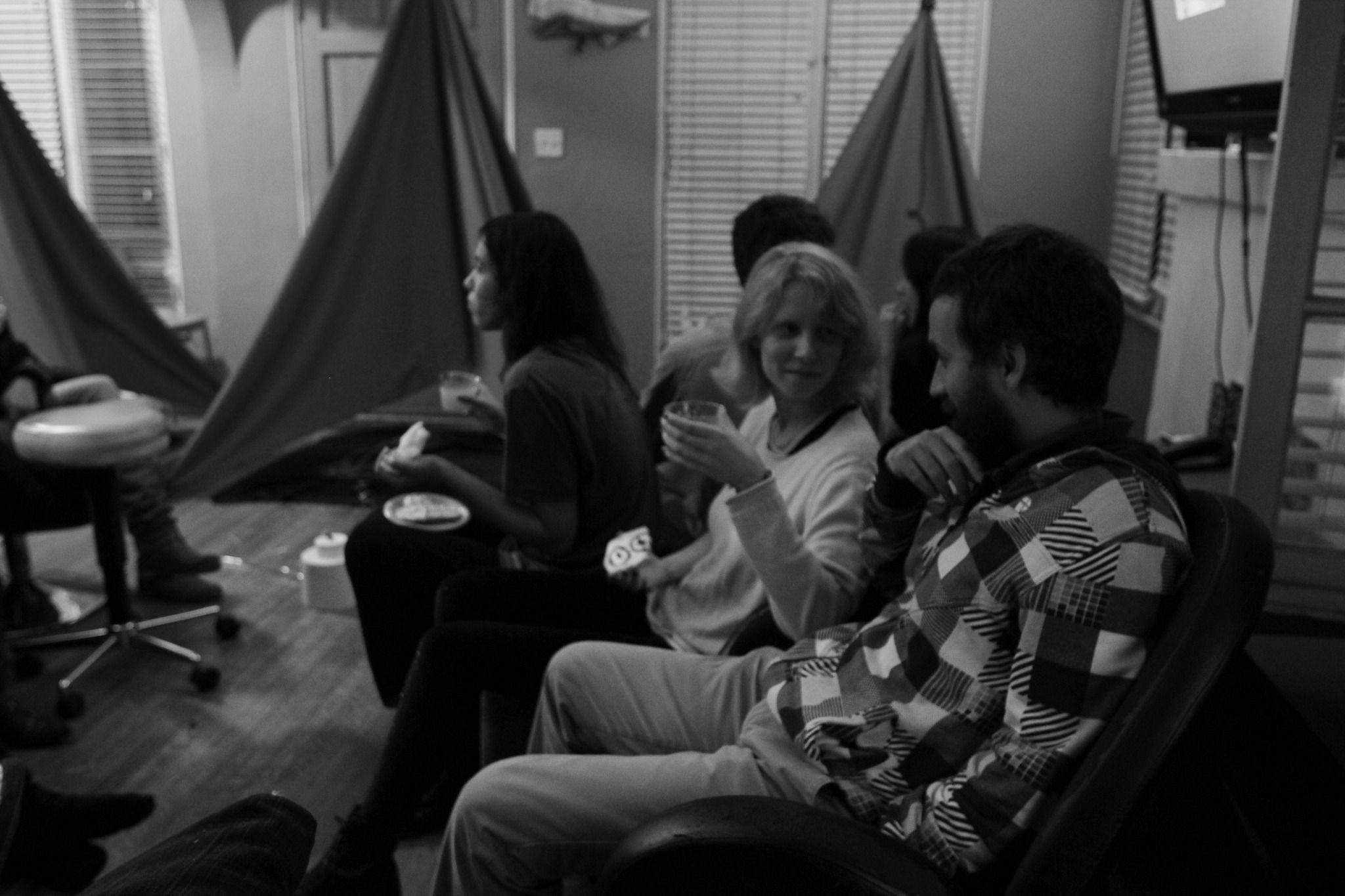 There is a dark, grayscale picture of various people sitting inside a small room. There are three tents in the background. There are three people sitting down and talking to each other. Some are holding cups. Everyone is smiling.