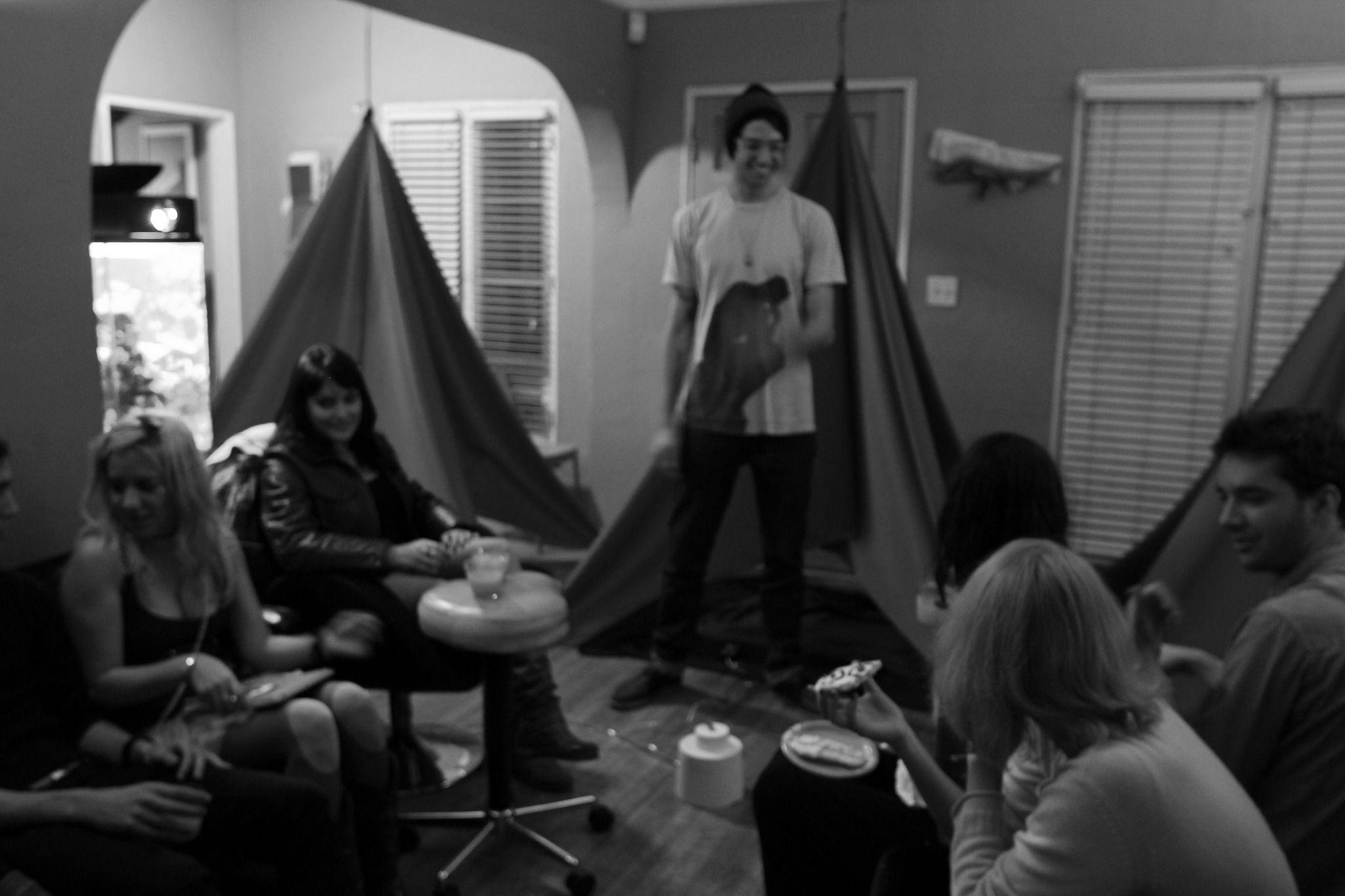 There is a dark, grayscale picture of various people sitting inside a small room. There are three tents in the background. One person is standing, and there are five or six people sitting down and talking to each other. Everyone is smiling.