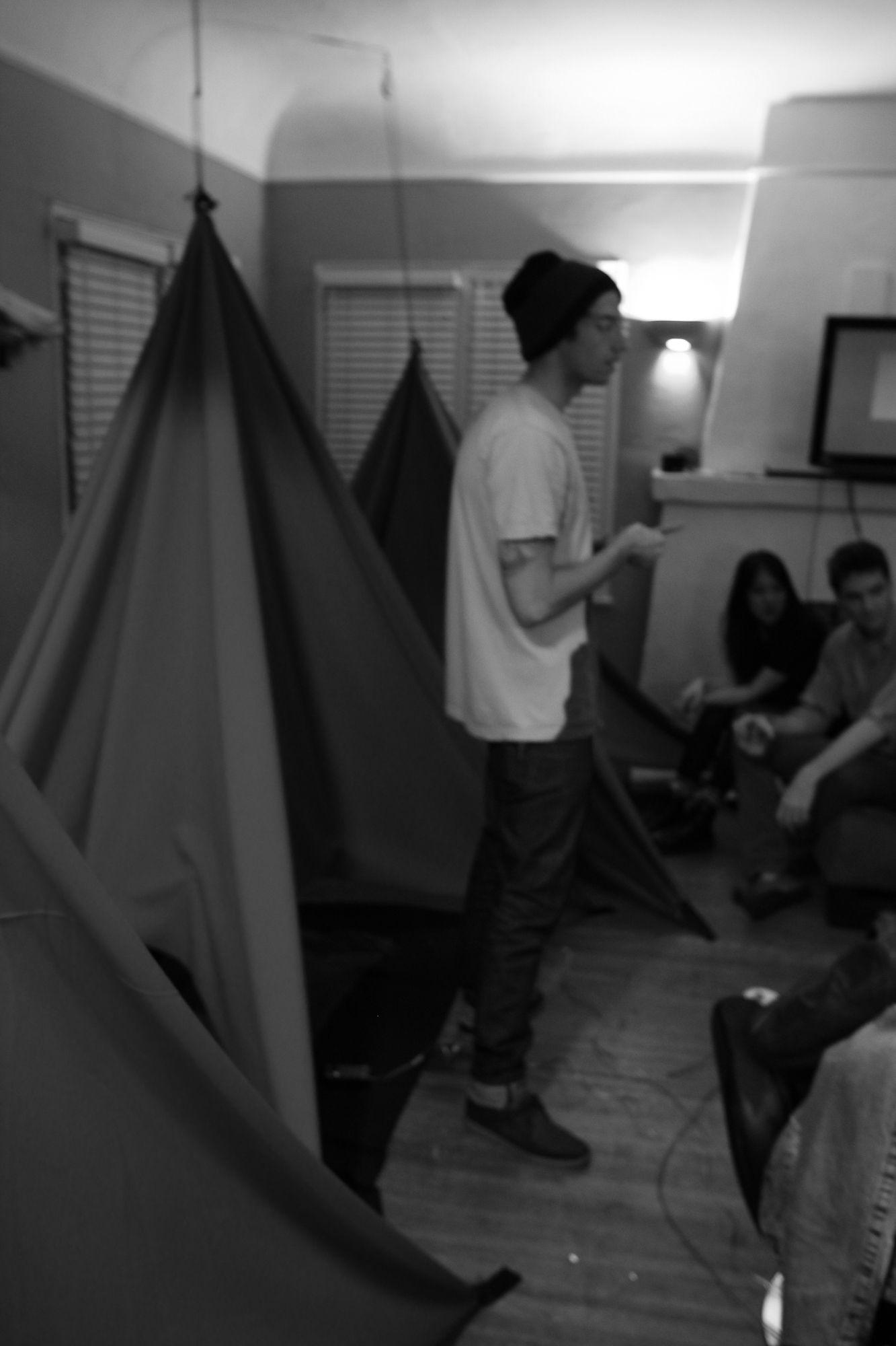 There is a dark, grayscale picture of people sitting inside a small room. There are three tents in the background. One person is standing and seems to be talking, and two people visible sitting down. Everyone is smiling.