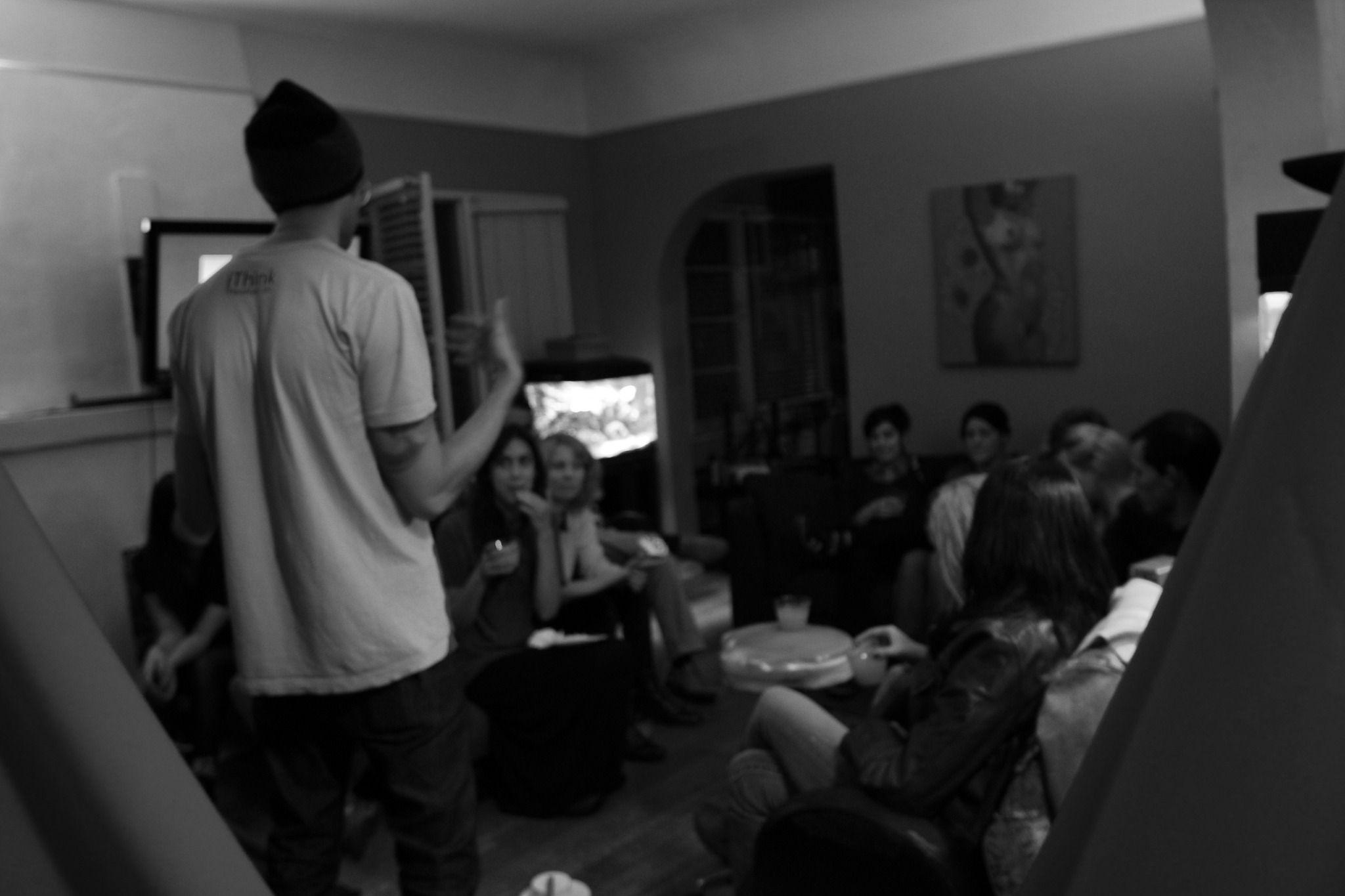 Someone is standing and talking to a large group of people who are sitting in a small room. The photo is dark and in grayscale. The person who is standing has their back to the camera.