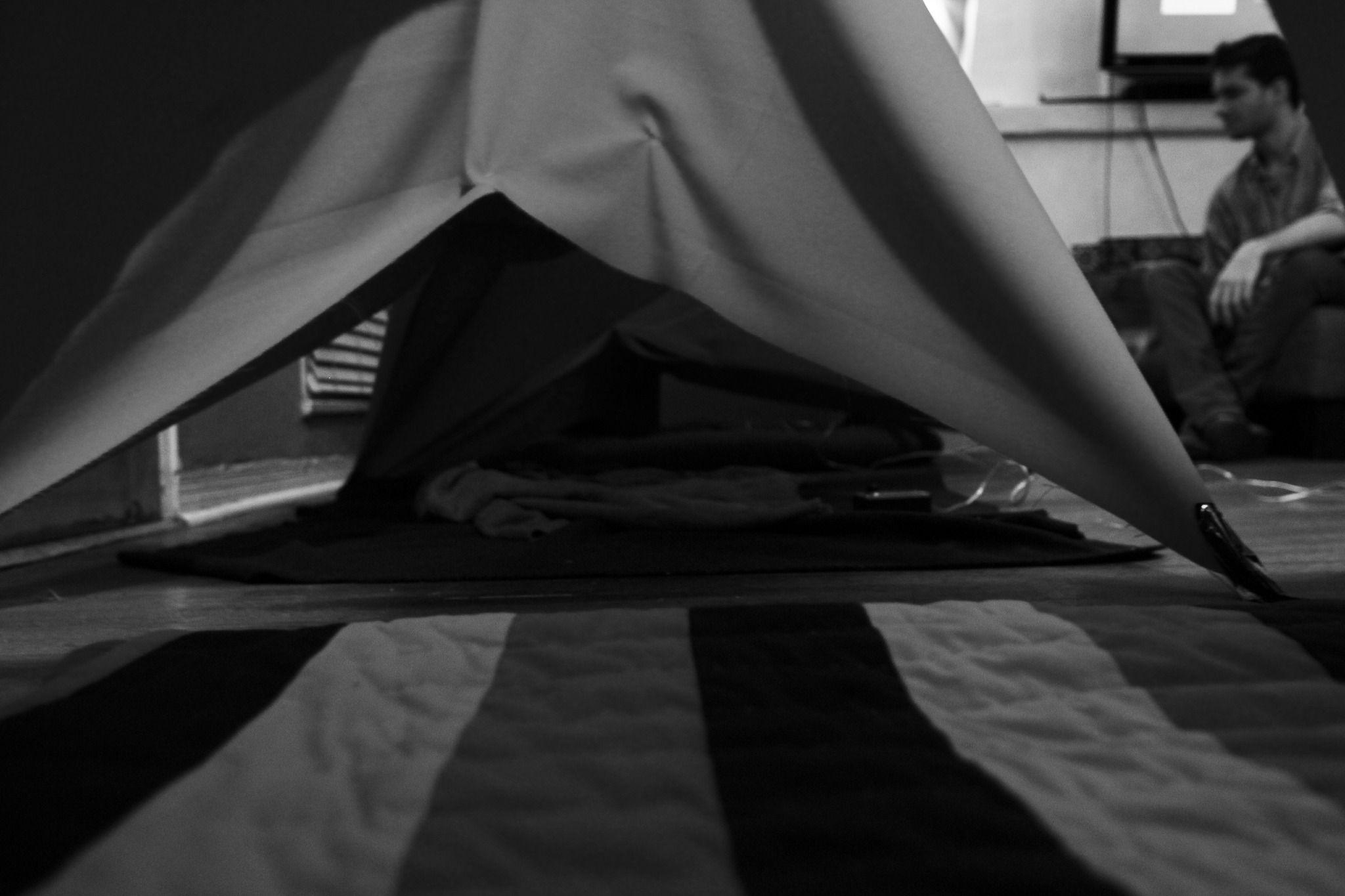 This is a photo possibly taken inside of a tent that is inside a small room. Things are grayscale and almost too dark to see, and a majority of the picture is obscured by the floor or the tent fabric. On the top right corner background there is a person t
