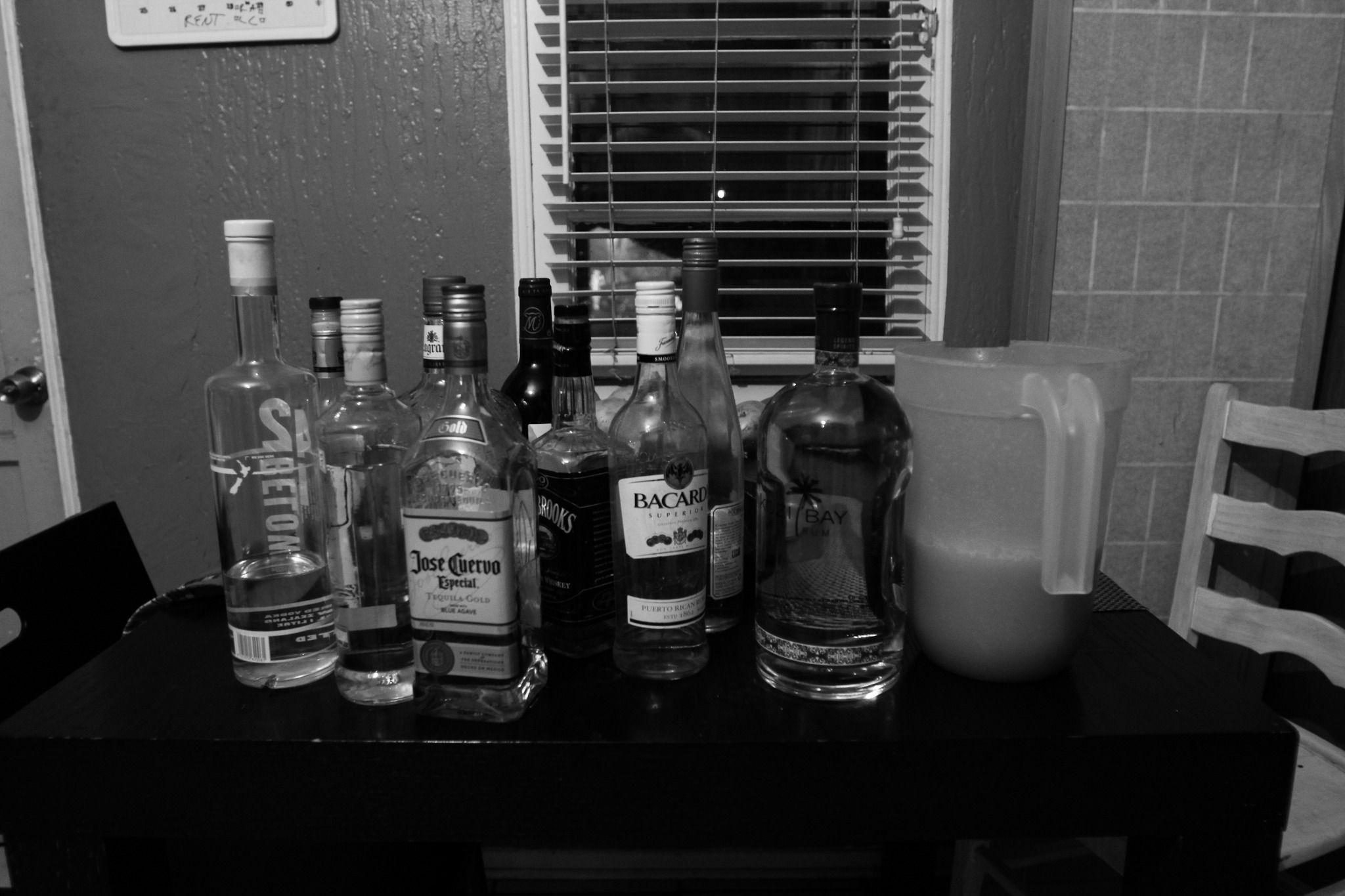 This is a grayscale dark photo of a small table with various bottles of alcohol. There is Jose Cuervo, Bacardi, 42 Below, and Jack Daniel's visible.